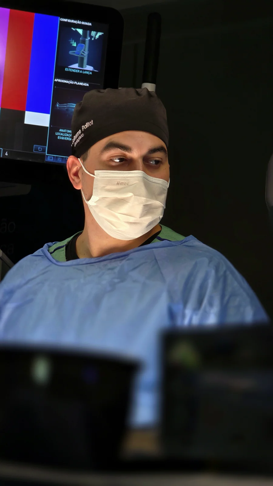 Dr. Guilherme Palitot em ambiente cirúrgico, com monitor do sistema robótico ao fundo.